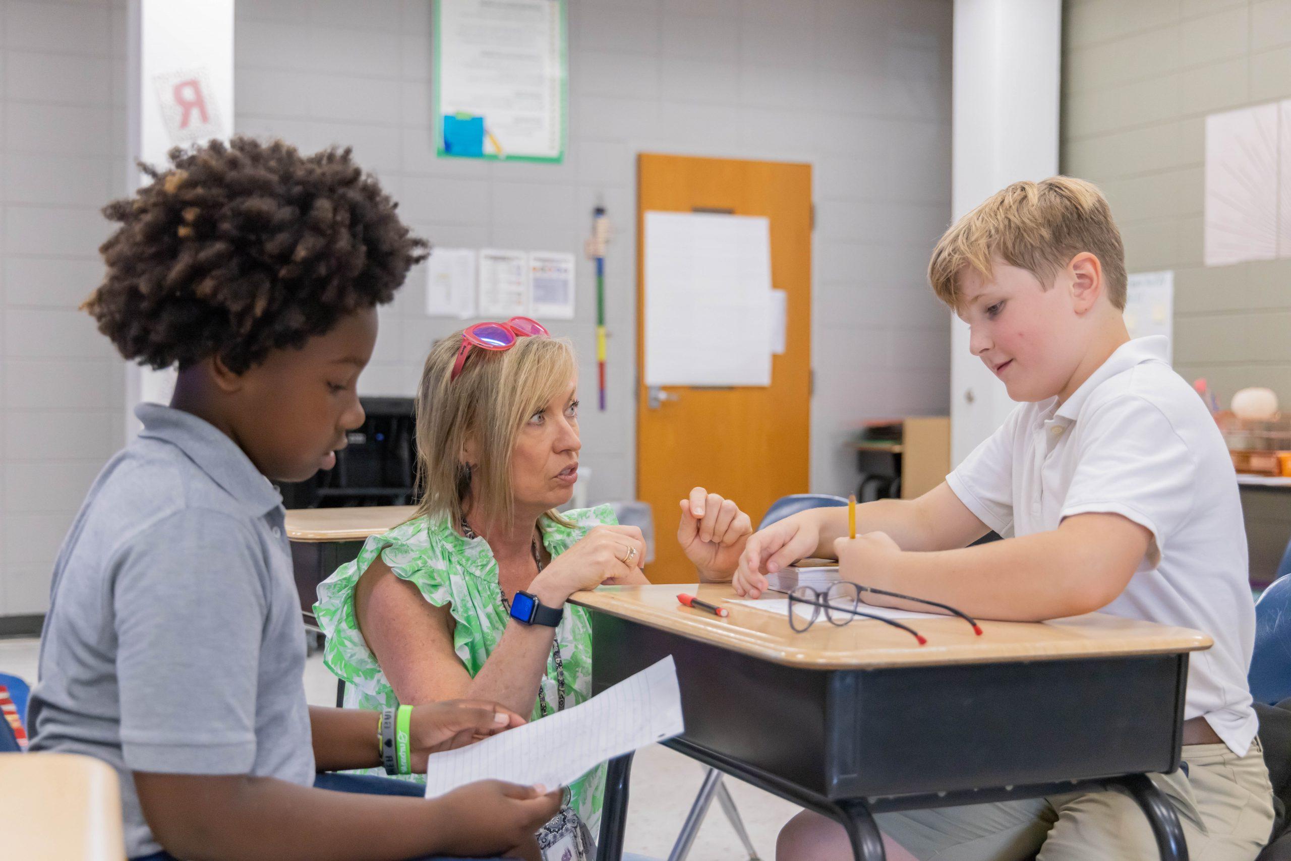 Two student grouped together at a desk with a teacher crouched down at the desk to help them with their class assignment.